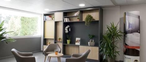 Una oficina moderna con tres sillas, una mesa pequeña, estanterías con libros y plantas, y un cartel grande en la pared.