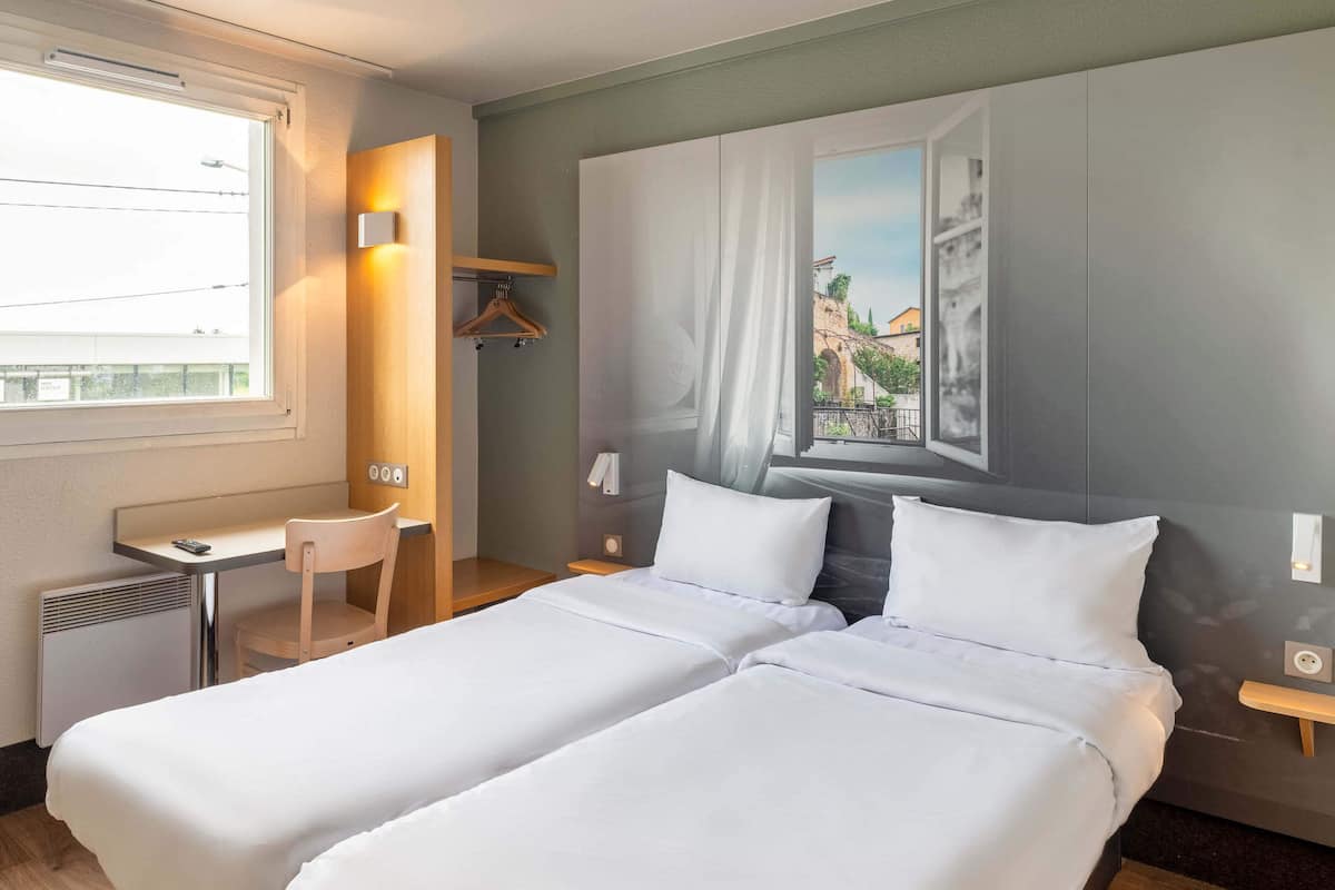 Habitación de hotel con dos camas individuales, un escritorio, una silla, un radiador, una ventana con vistas a edificios e iluminación empotrada en la pared.