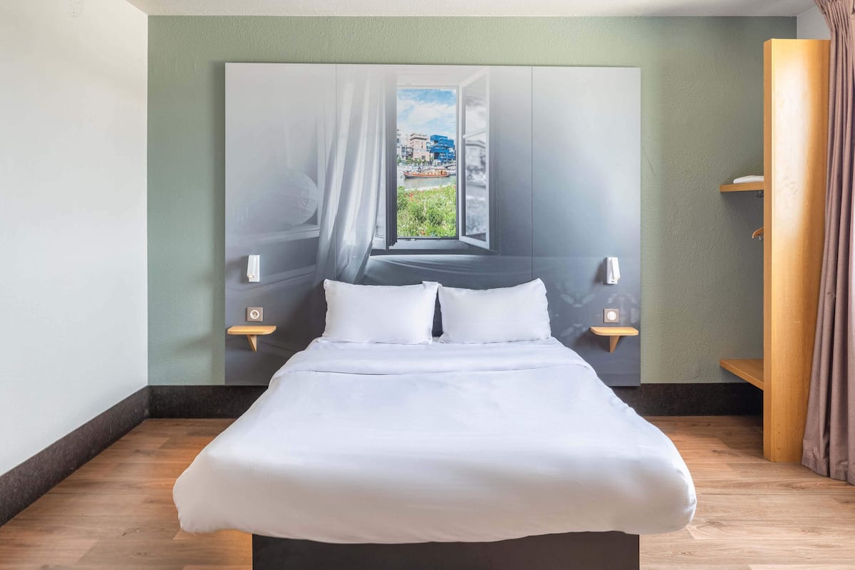 Una habitación de hotel moderna con una cama grande, vista de un paisaje urbano a través de una ventana abierta y una mesita de noche de madera con una lámpara.