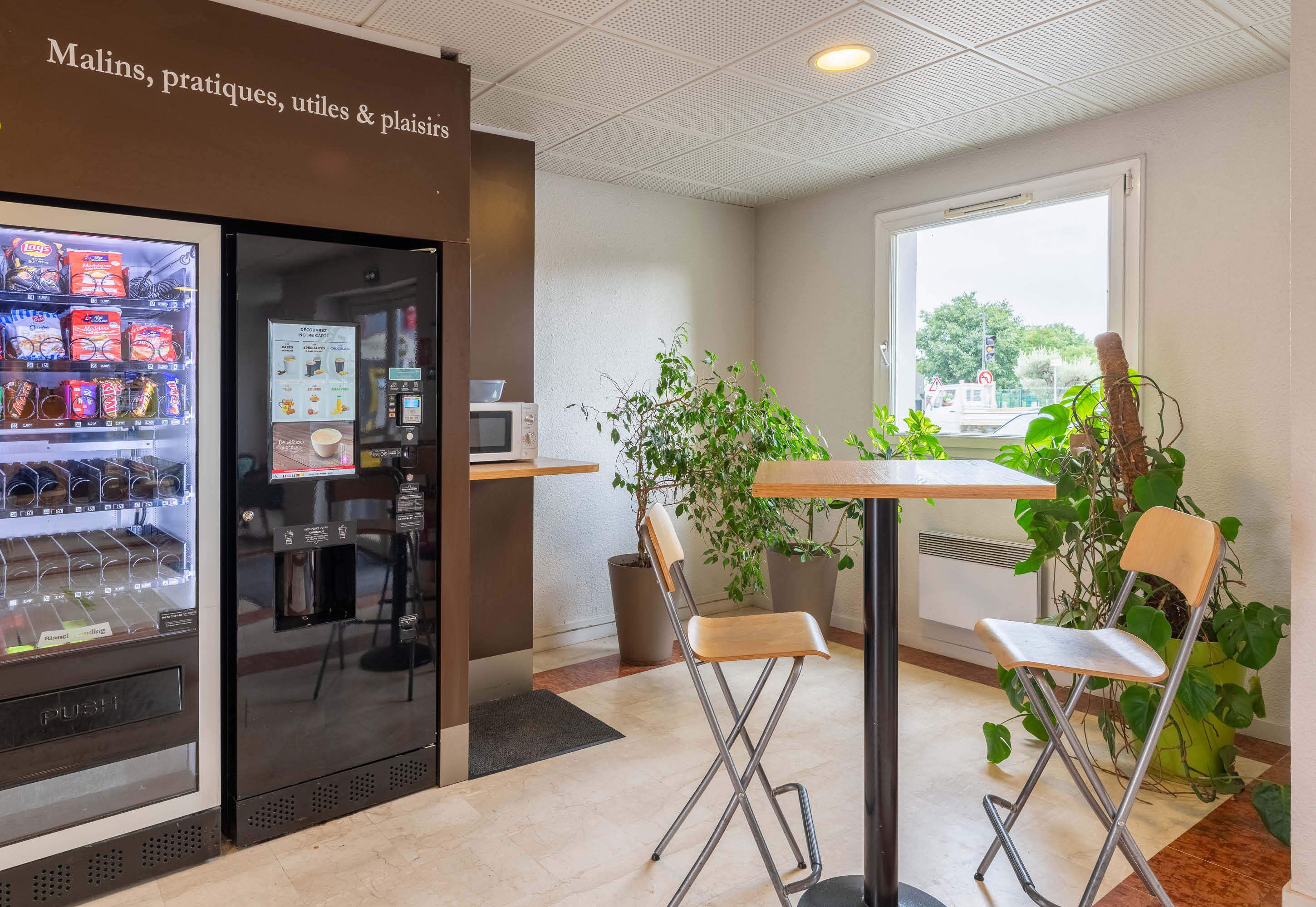 Un café au décor intérieur propre et moderne, avec un distributeur automatique, un espace salon et des plantes en pot.