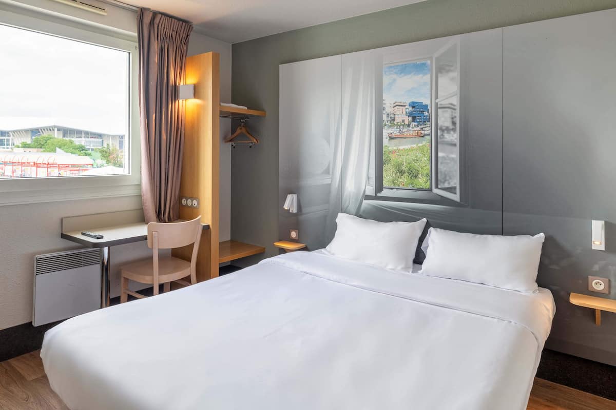 Habitación de hotel con una cama grande, un escritorio con silla, una mesita y una ventana con cortinas.