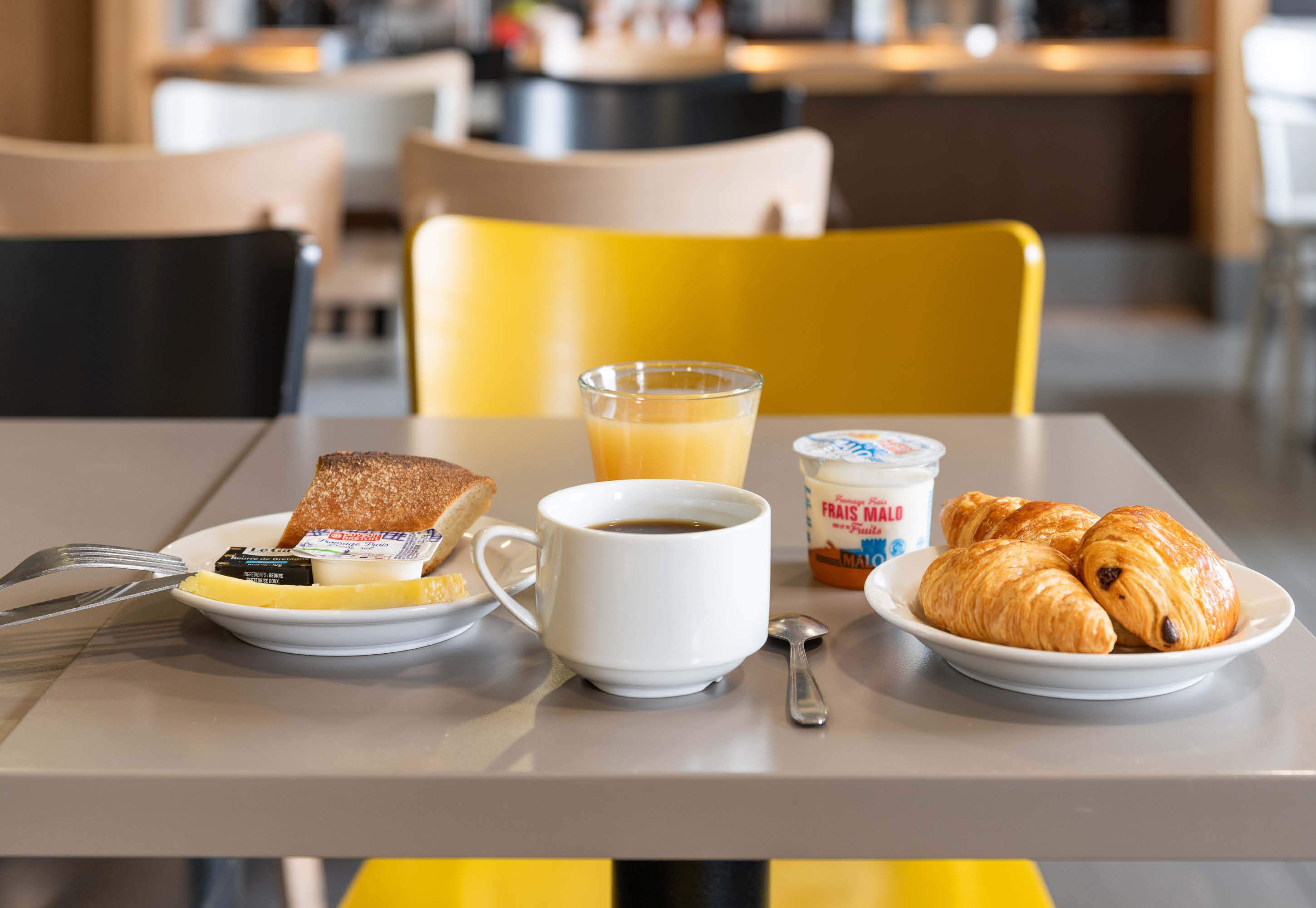 A breakfast setting with toast, a cup of coffee, a glass of orange juice, yogurt, and croissants on a table.