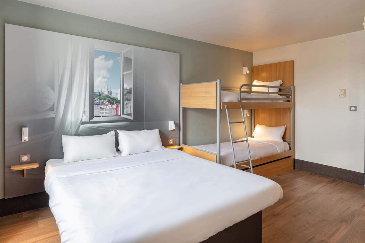 Una habitación de hotel con una cama grande, una litera, un ventanal con vista a la ciudad y un estante de pared.