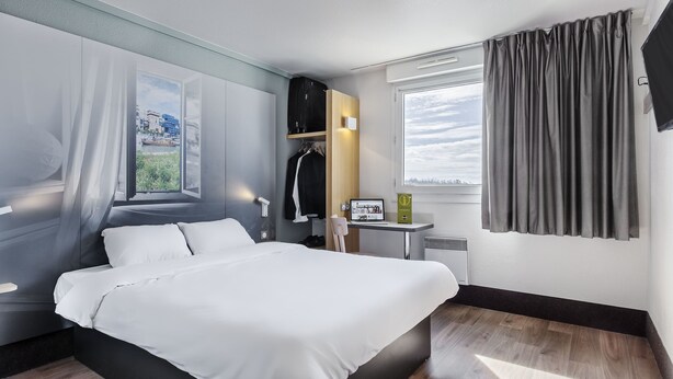 Een moderne hotelkamer met een groot bed, een klein tafeltje met een laptop, een kapstok en uitzicht op de stad door het raam.