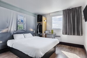 Een moderne hotelkamer met een groot bed, een klein tafeltje met een laptop, een kapstok en uitzicht op de stad door het raam.