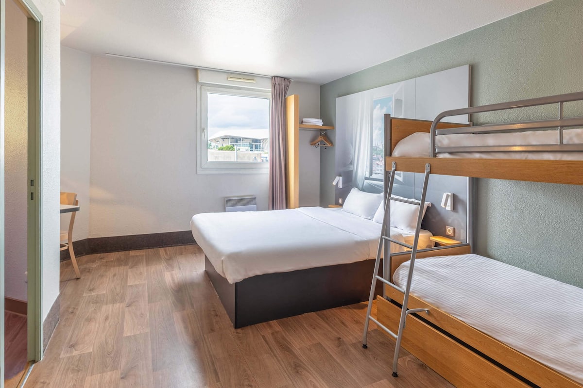Habitación de hotel con literas, una cama individual, una ventana con cortinas, suelo de madera y vistas a edificios.