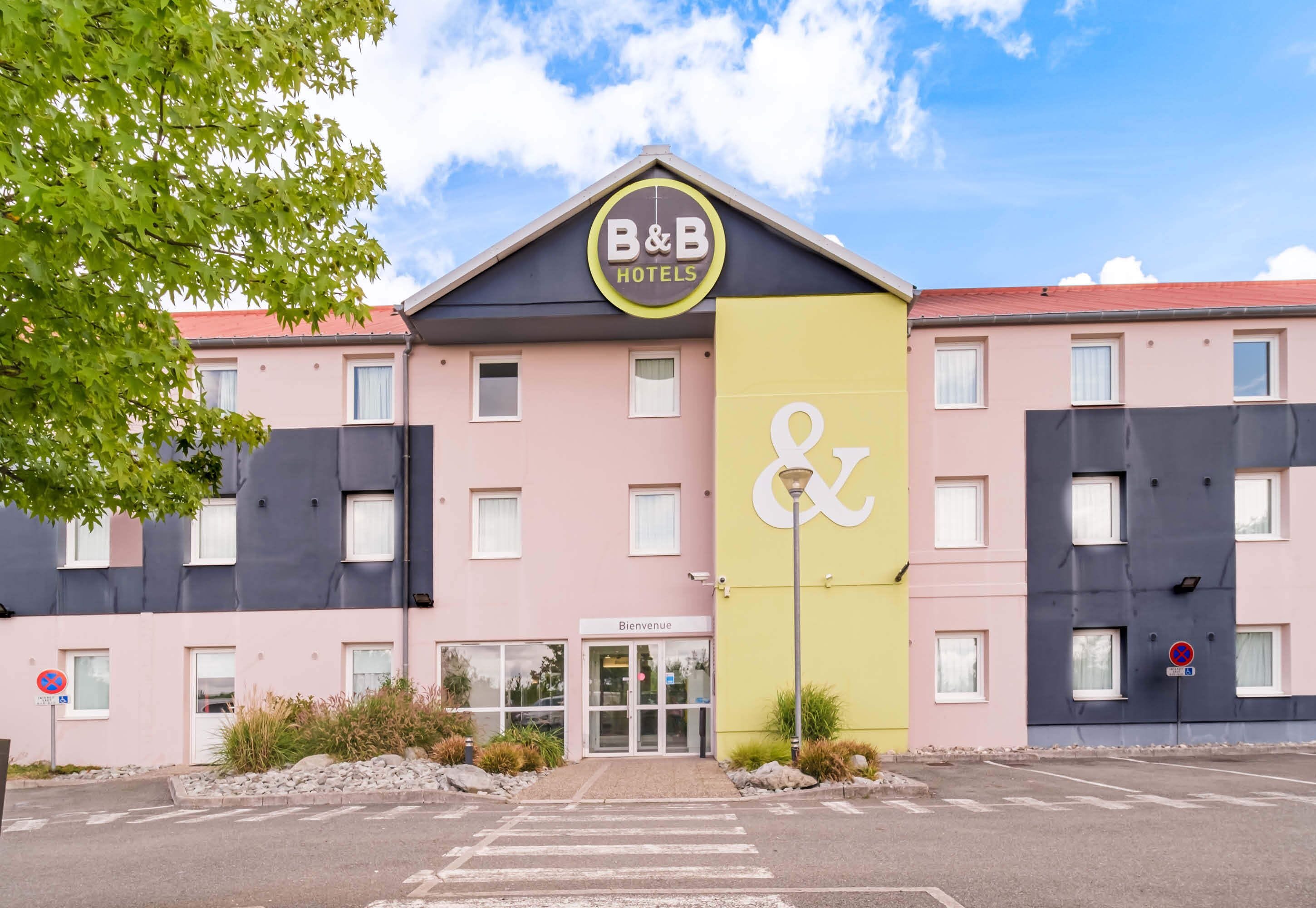 Un hotel B&B con fachada rosa, un letrero vertical amarillo y una entrada con el rótulo 'Bienvenue'.