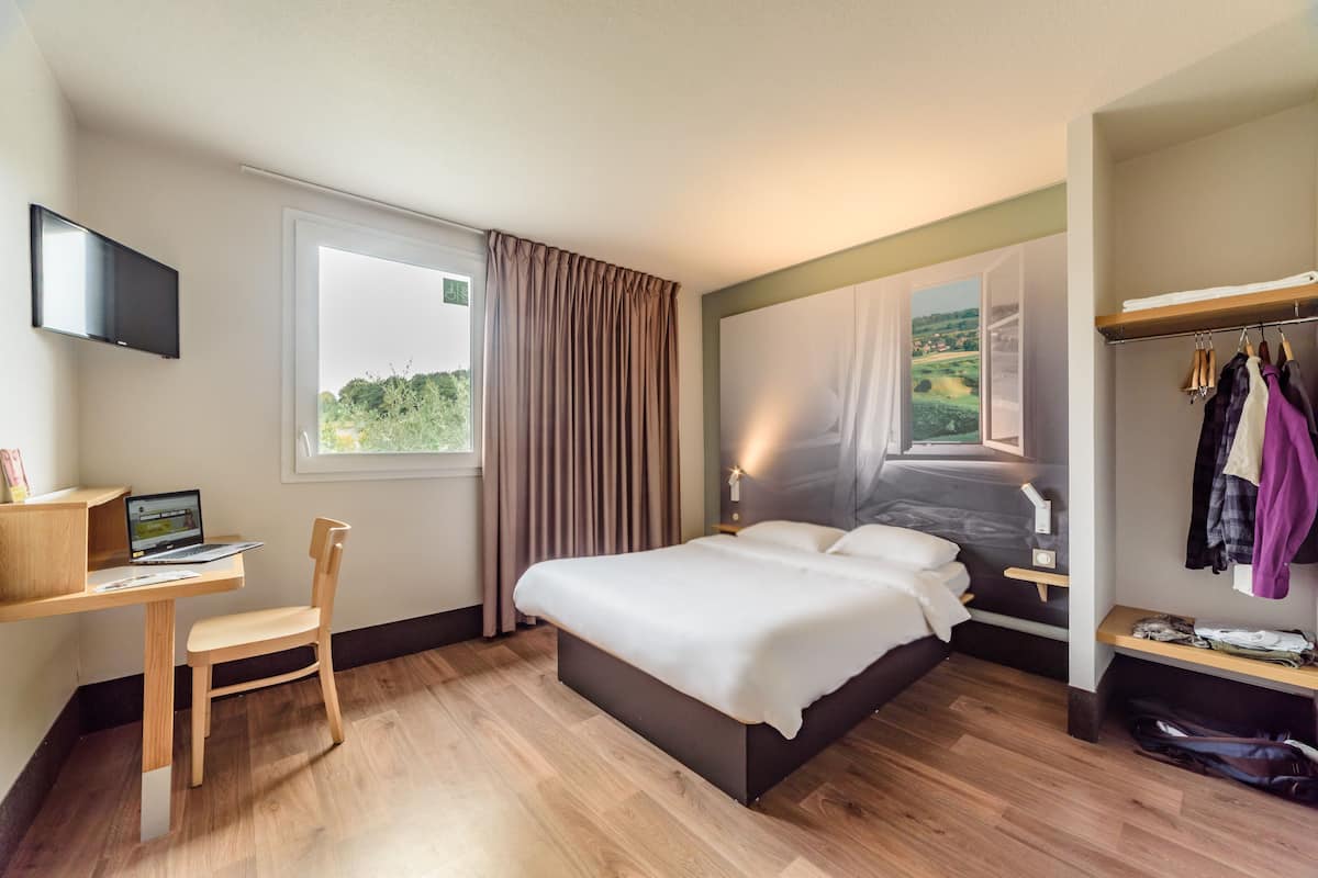 A hotel room with a bed, a desk with a laptop, a chair, a TV mounted on the wall, and a window with curtains.