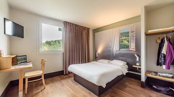 Habitación de hotel con cama, escritorio con una laptop, una silla, televisor montado en la pared y ventana con cortinas.