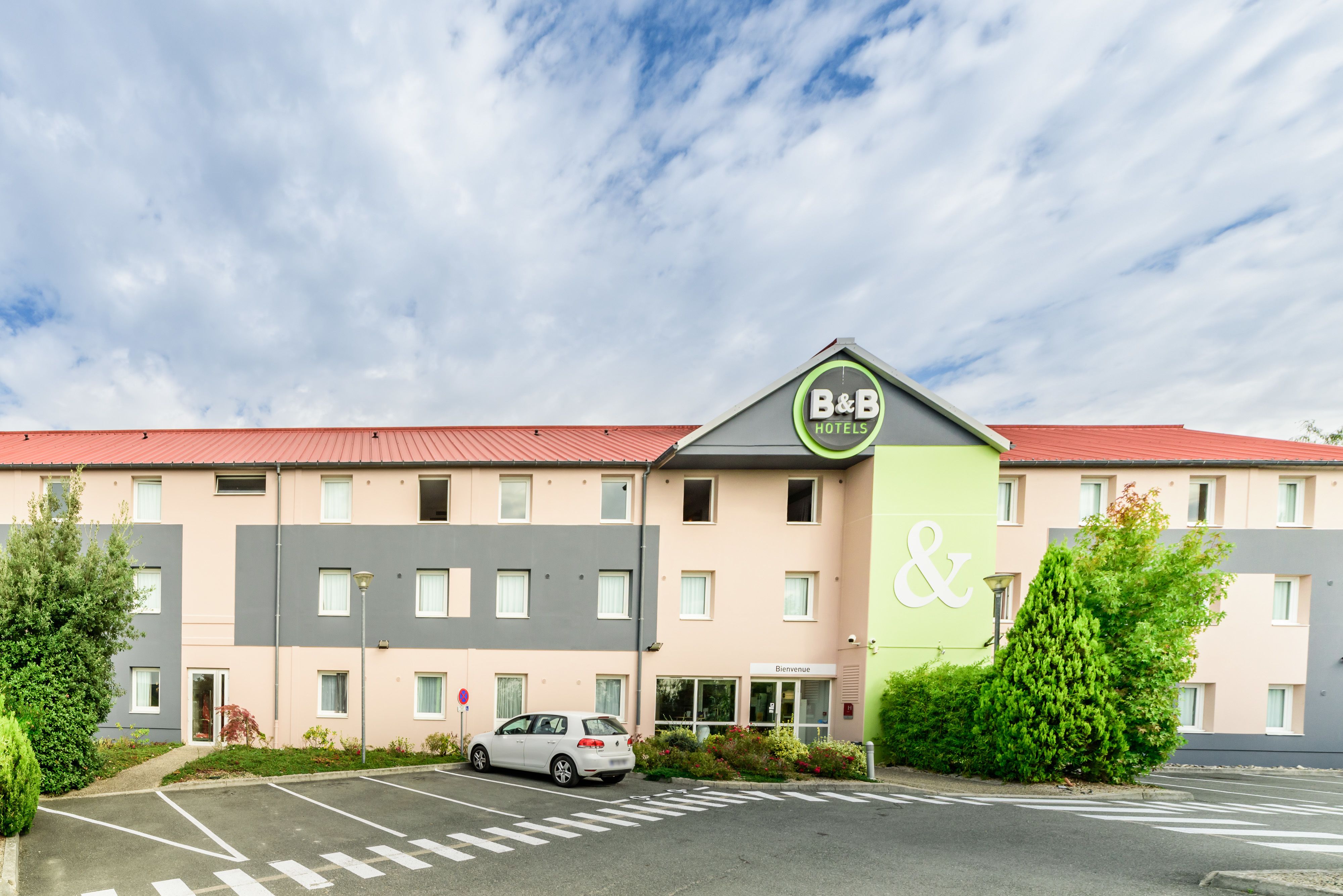 A B&B hotel with a red roof, a green and grey facade, and a parking lot with a white car.