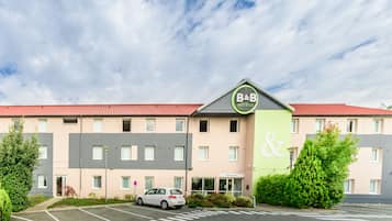 A B&B hotel with a red roof, a green and grey facade, and a parking lot with a white car.