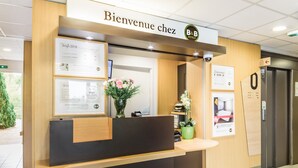 A reception desk with a sign reading "Bienvenue chez B&B Hotels," a framed menu, and a potted plant. - B&B HOTEL Belfort (Belfort)