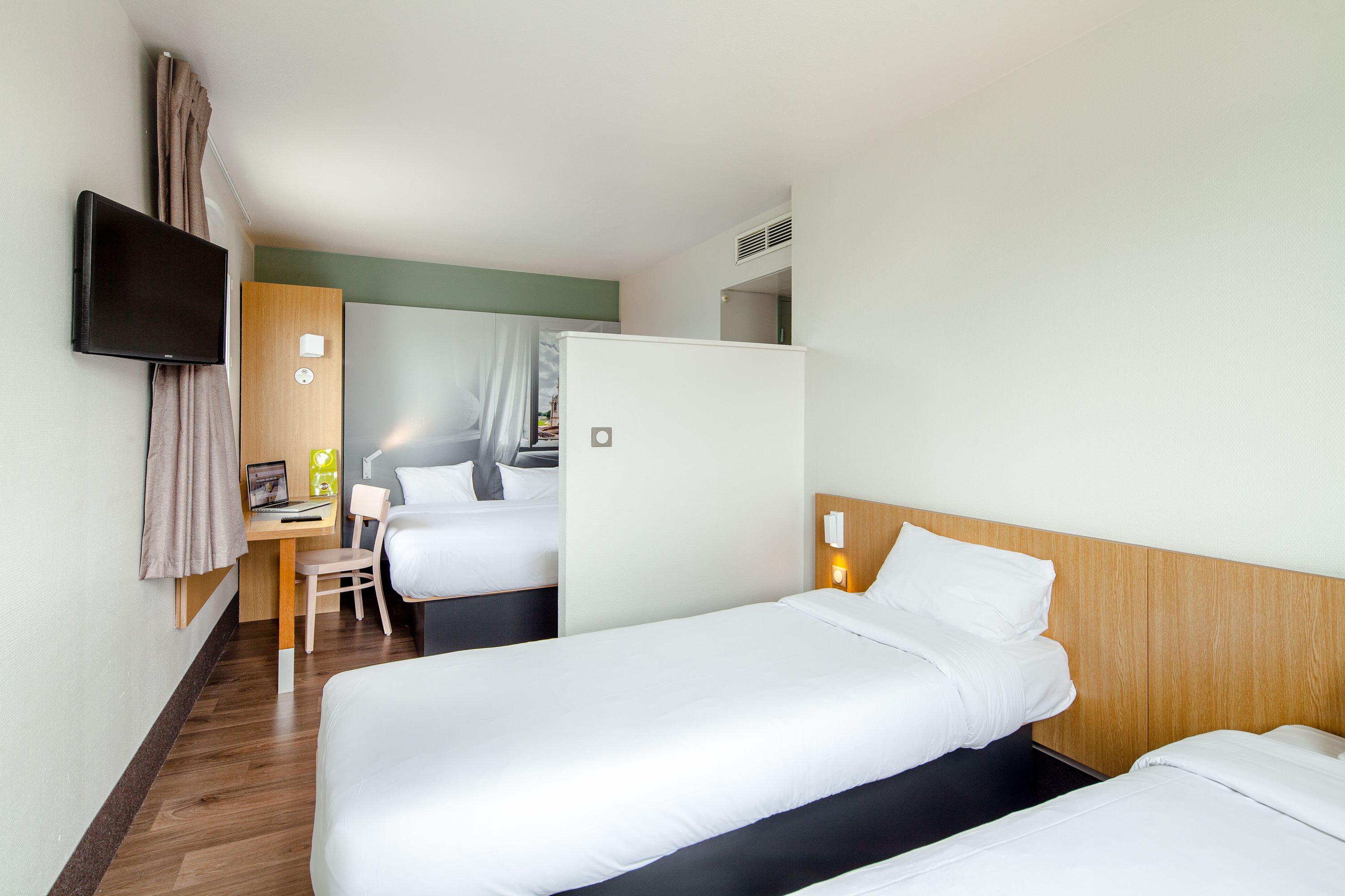 Habitación de hotel con dos camas individuales, un escritorio con silla, un televisor de pantalla plana en la pared y un refrigerador pequeño.