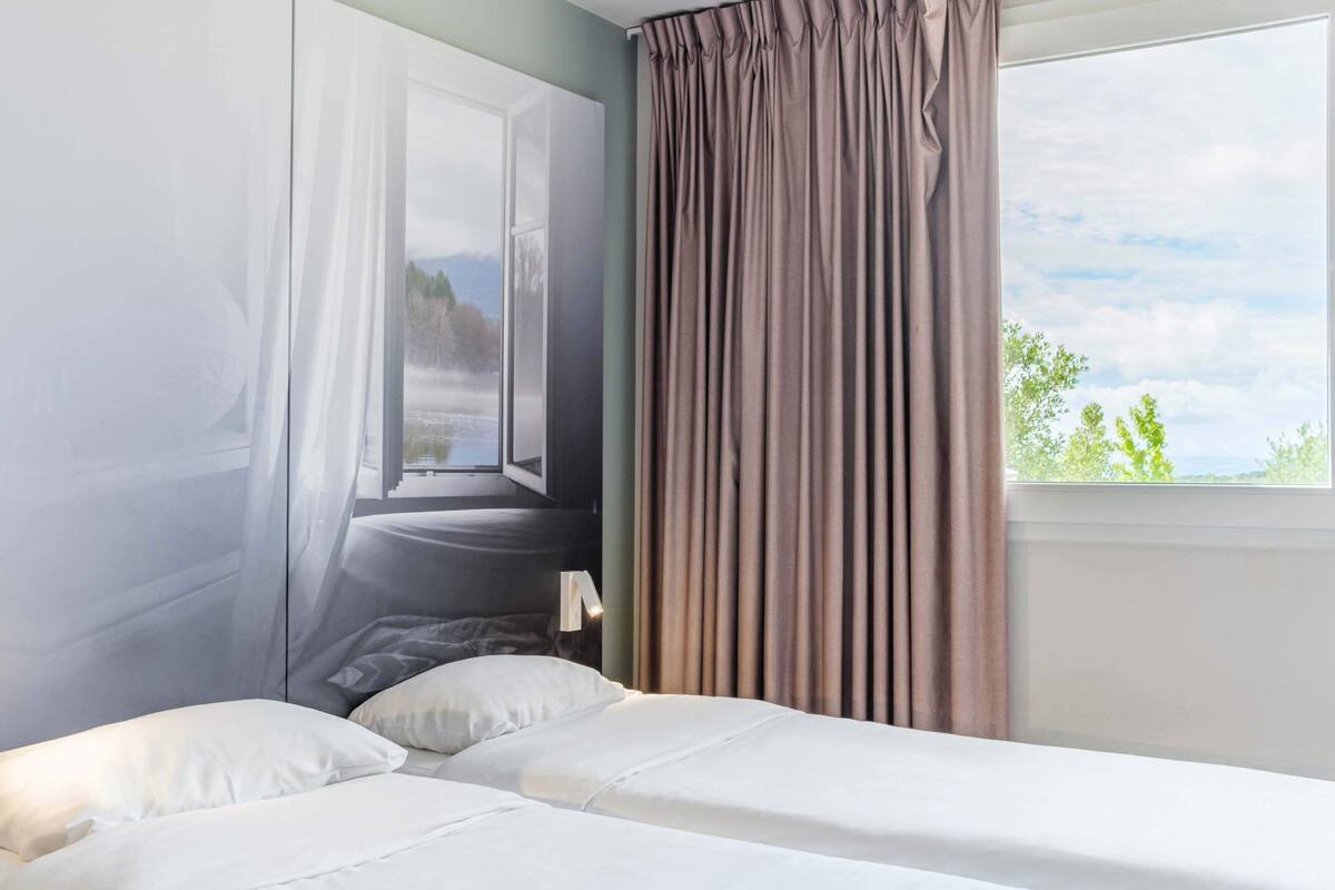 A hotel room with a bed, a window with curtains, and a view of a landscape.
