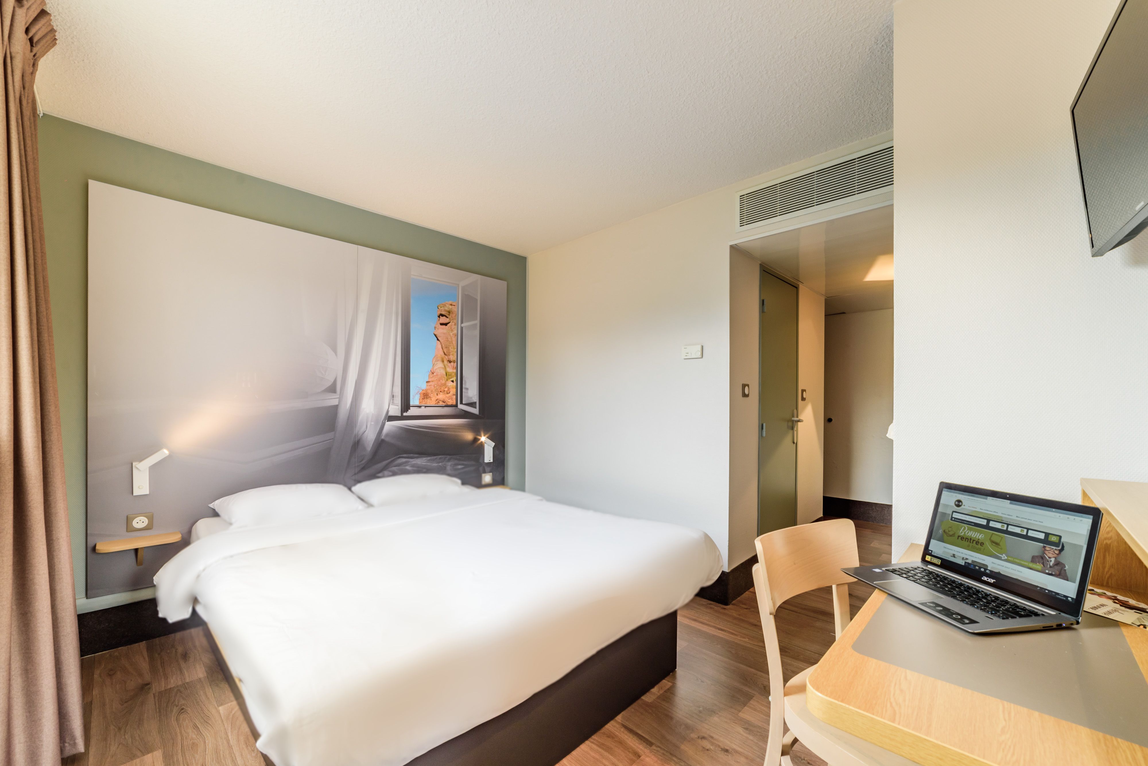 Habitación de hotel con una cama, un escritorio con una computadora portátil, una silla y una puerta que da a otra habitación.