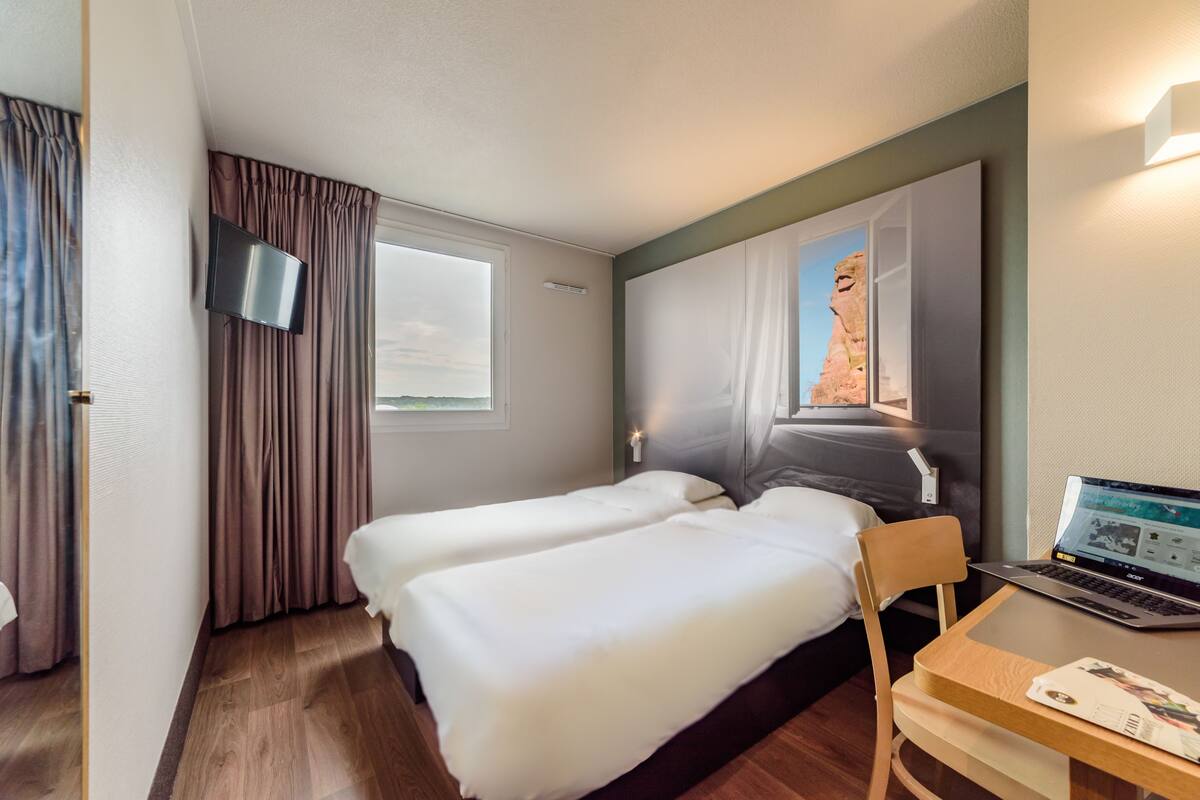 A hotel room with two beds, a desk with a laptop, a TV, and a window with curtains.