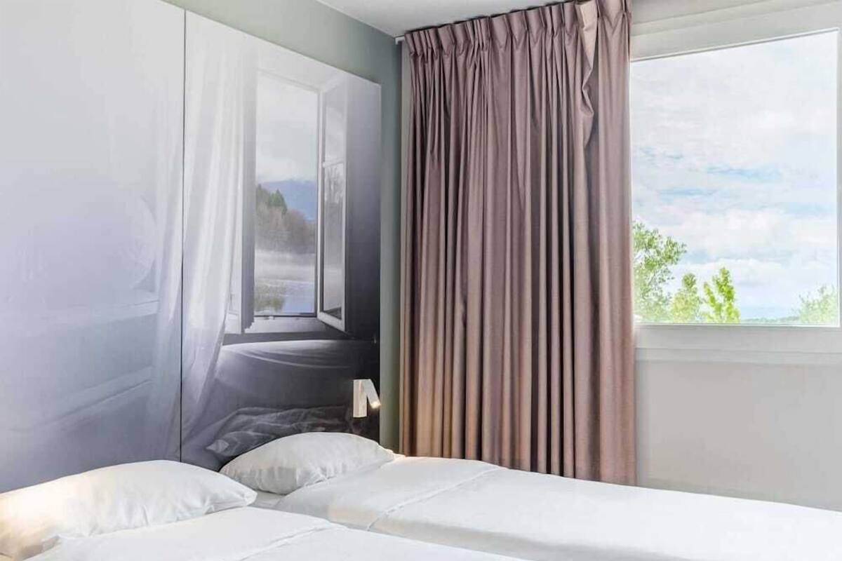 A hotel room with a bed, a window with curtains, and a view of the outdoors.
