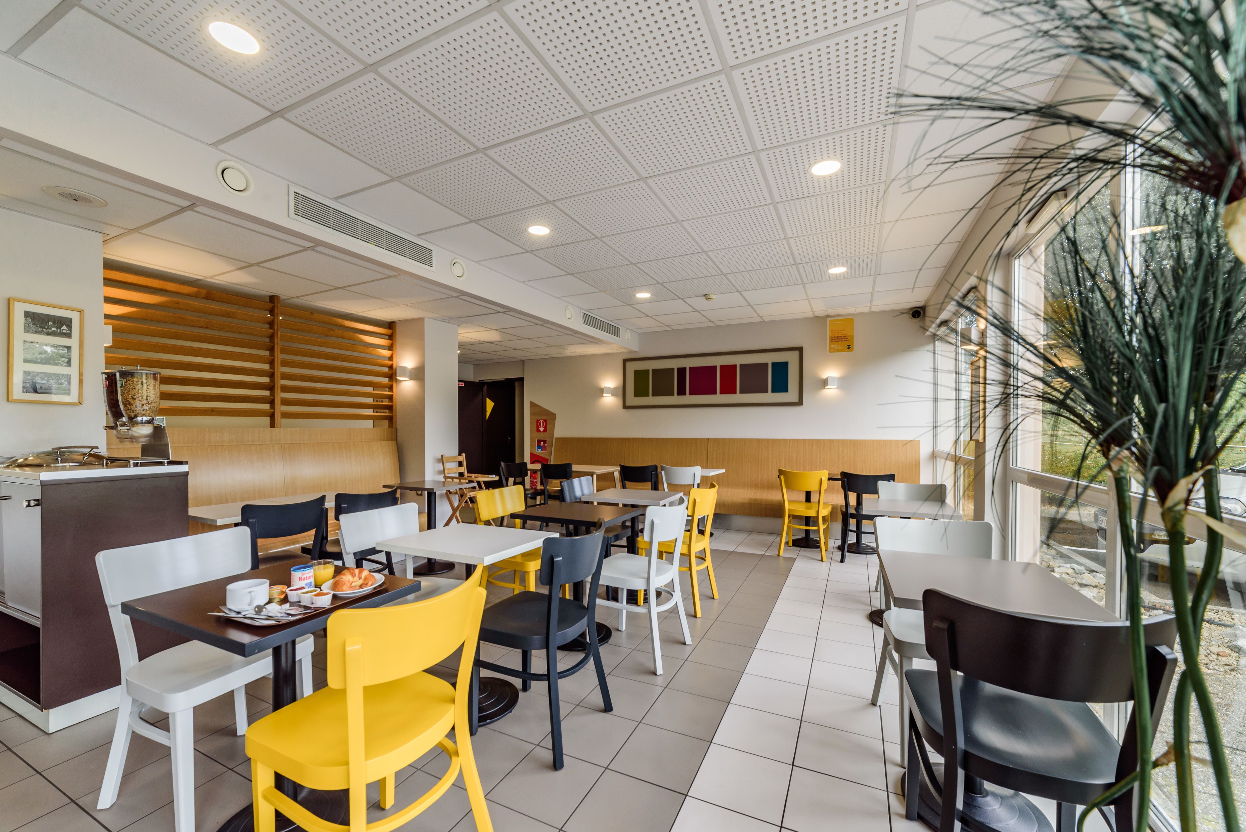 A restaurant with a modern interior, featuring a mix of white and yellow chairs, a central counter with a menu display, and a large window providing natural light.