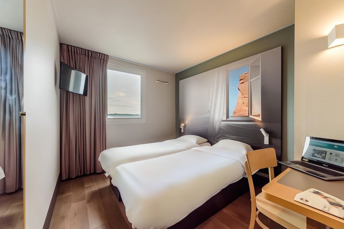 A hotel room with two beds, a desk with a laptop, a TV, and a window with curtains.