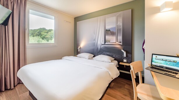 A hotel room with a bed, a laptop on a desk, and a window with curtains. - B&B HOTEL Belfort (Belfort)