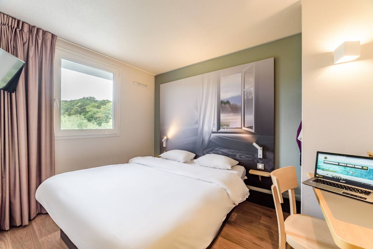 A hotel room with a bed, a desk with a laptop, a window with curtains, and a wall-mounted light.