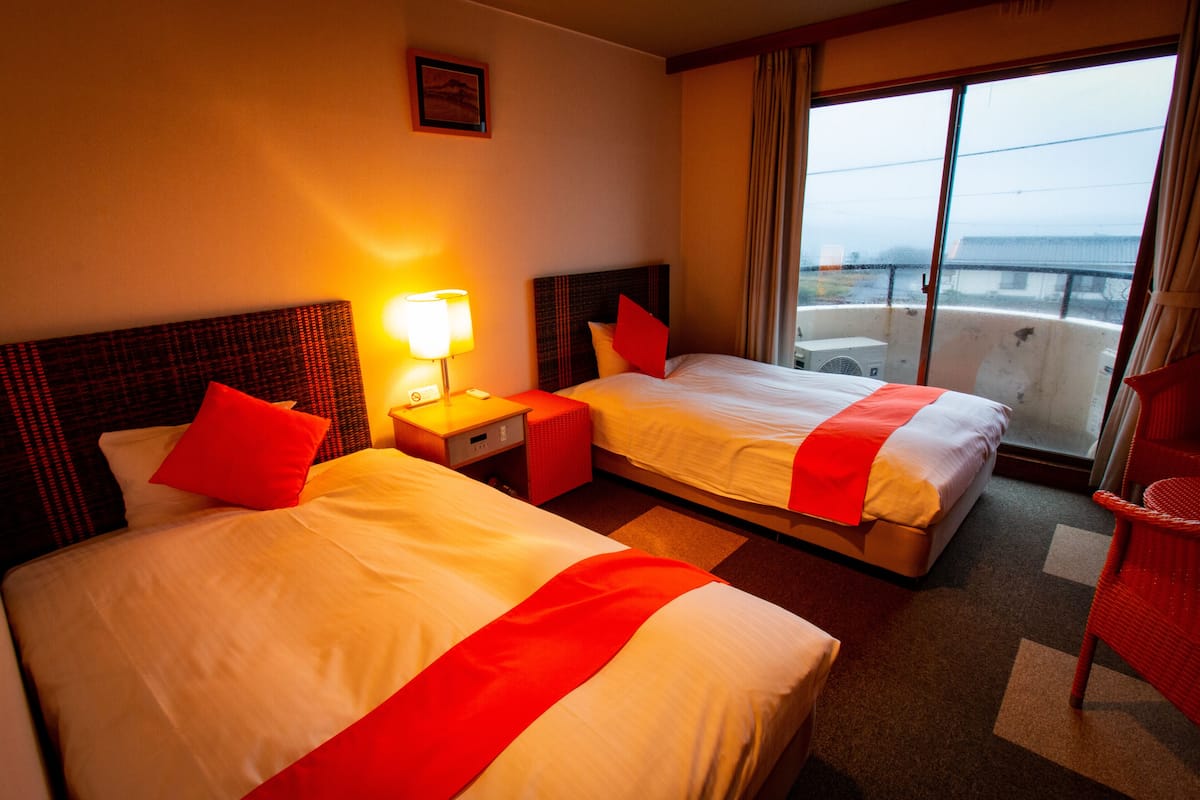 A hotel room with two beds, a nightstand with a lamp, a window with a view of the city, and a red chair.