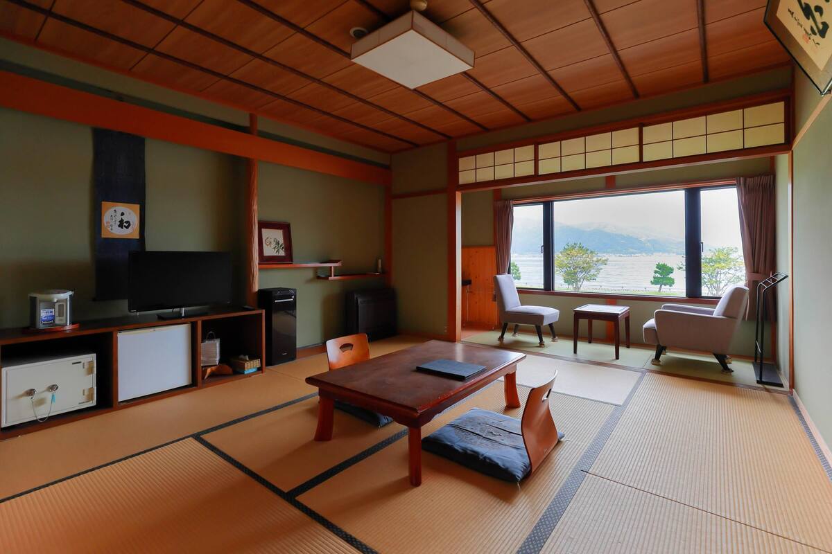 A traditional Japanese room with tatami flooring, a low table, chairs, a television, and a view of a body of water.