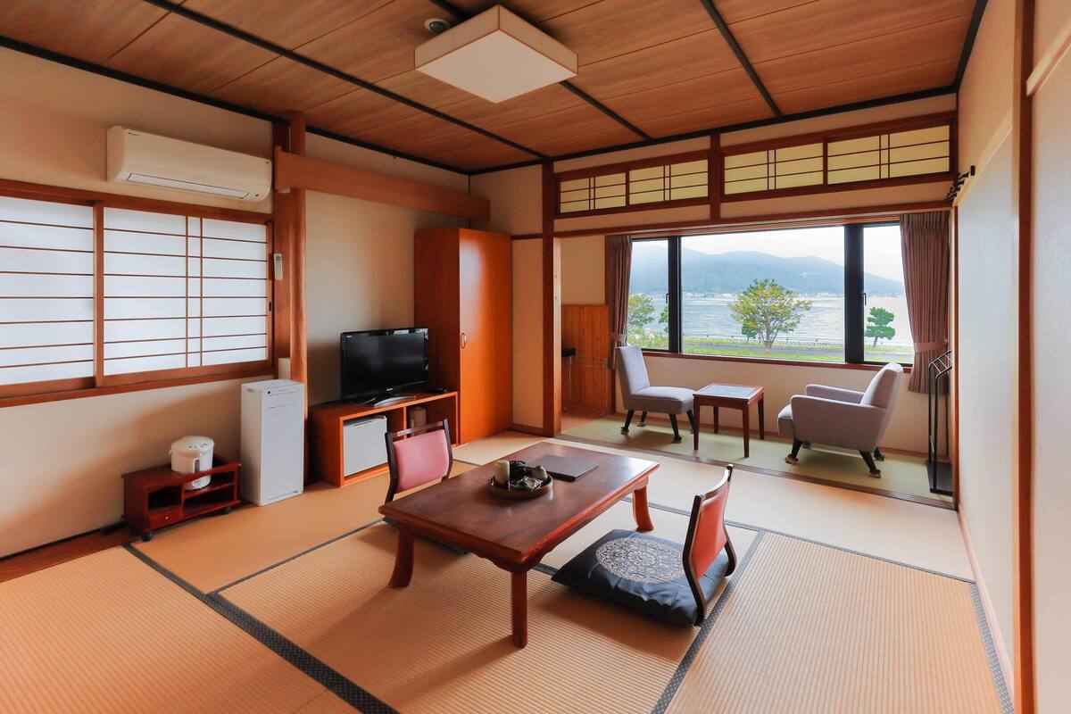 A traditional Japanese room with tatami flooring, a television, a small refrigerator, a wooden table, and a view of a lake.