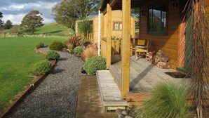 A wooden cabin with a covered porch, a gravel pathway, and a fenced yard with various plants. - The River Lodge (Ohakune)