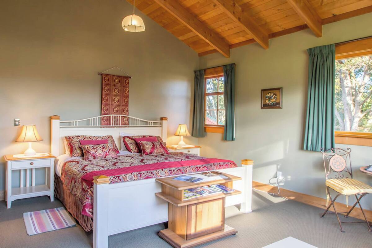 Un dormitorio con una cama de madera, dos mesitas de noche con lámparas, una pequeña mesita auxiliar de madera, una silla y una ventana con cortinas.