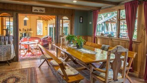 A wooden dining area with a large table and benches, surrounded by windows with curtains. - The River Lodge (Ohakune)