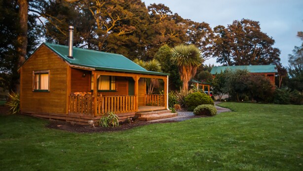 A wooden cabin with a green roof, surrounded by a well-maintained lawn and various trees. - The River Lodge (Ohakune)