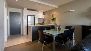 A modern kitchen with a central island, a dining area with a glass table and black chairs, and a black leather sofa. - ABC Accommodation - Spencer Street (Melbourne)