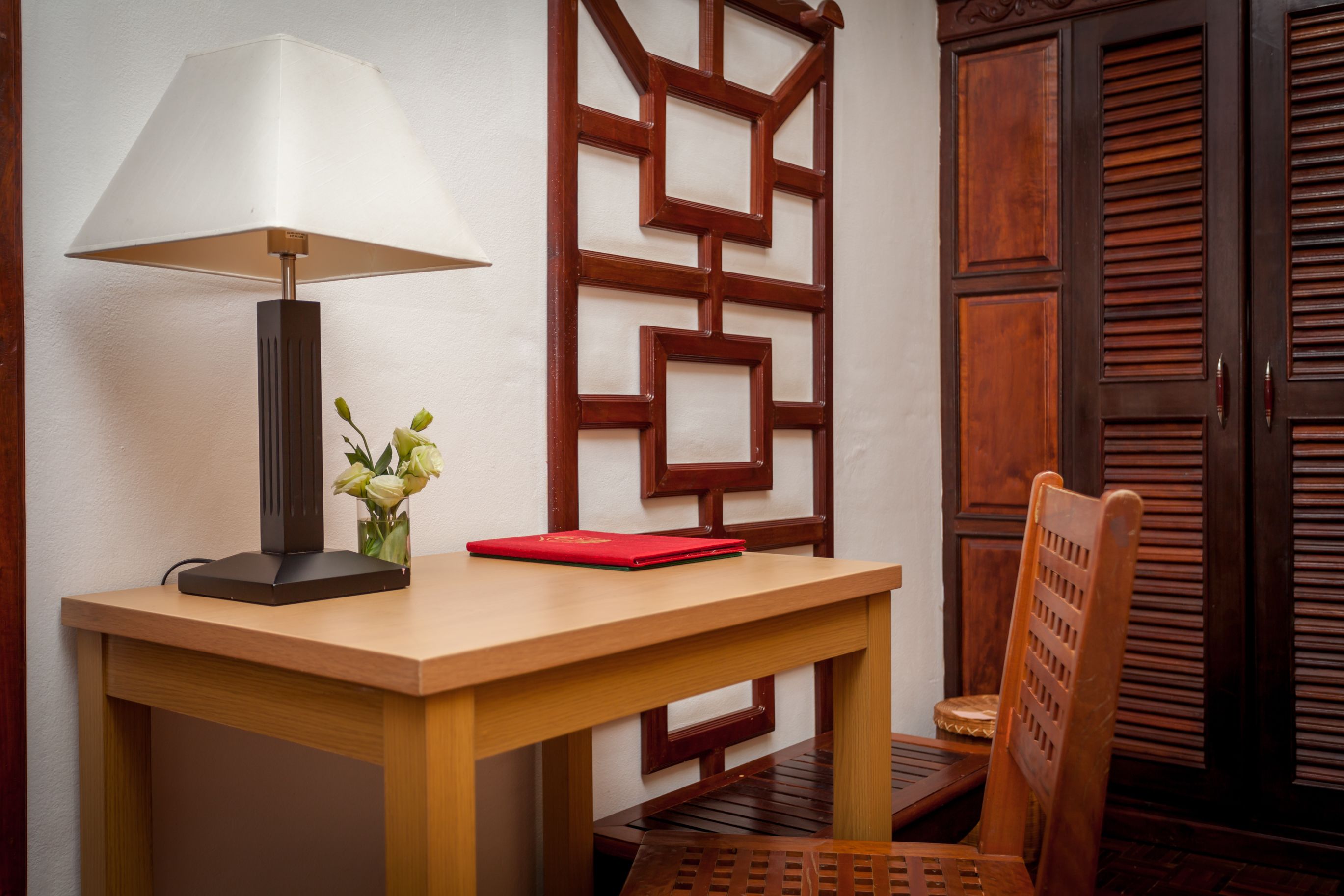 A wooden table with a lamp, a red notebook, and a vase with flowers.