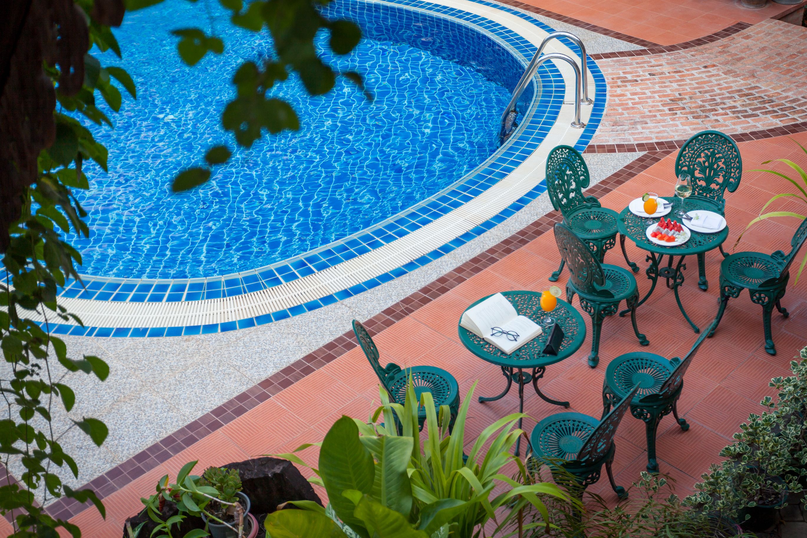 A pool area with a tiled edge, surrounded by green metal chairs and tables, and potted plants.