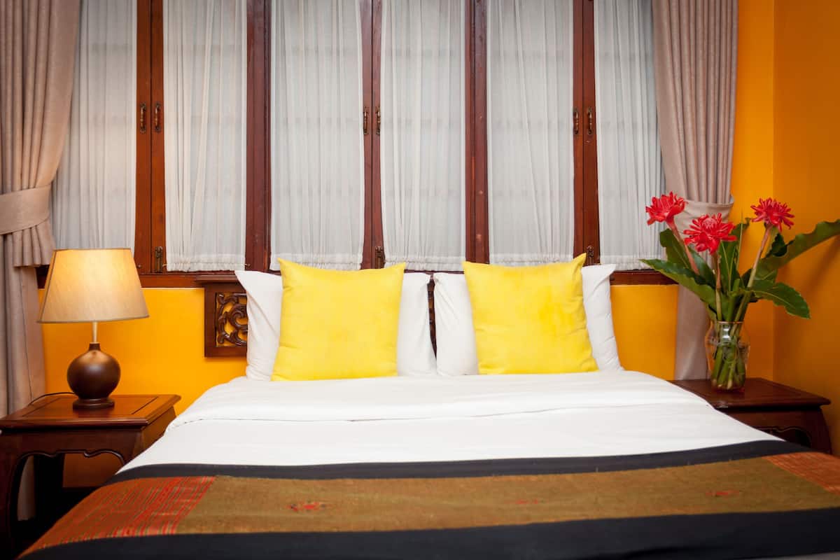 Una habitación de hotel con una cama, almohadones amarillos, una lámpara de noche, un jarrón con flores y cortinas.