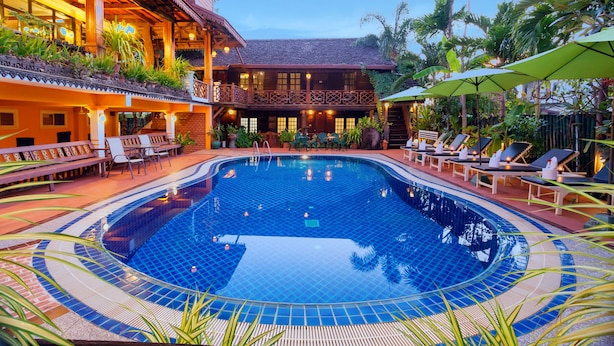 Una piscina con sillas de descanso y sombrillas, rodeada de plantas tropicales y un edificio de madera con balcones.