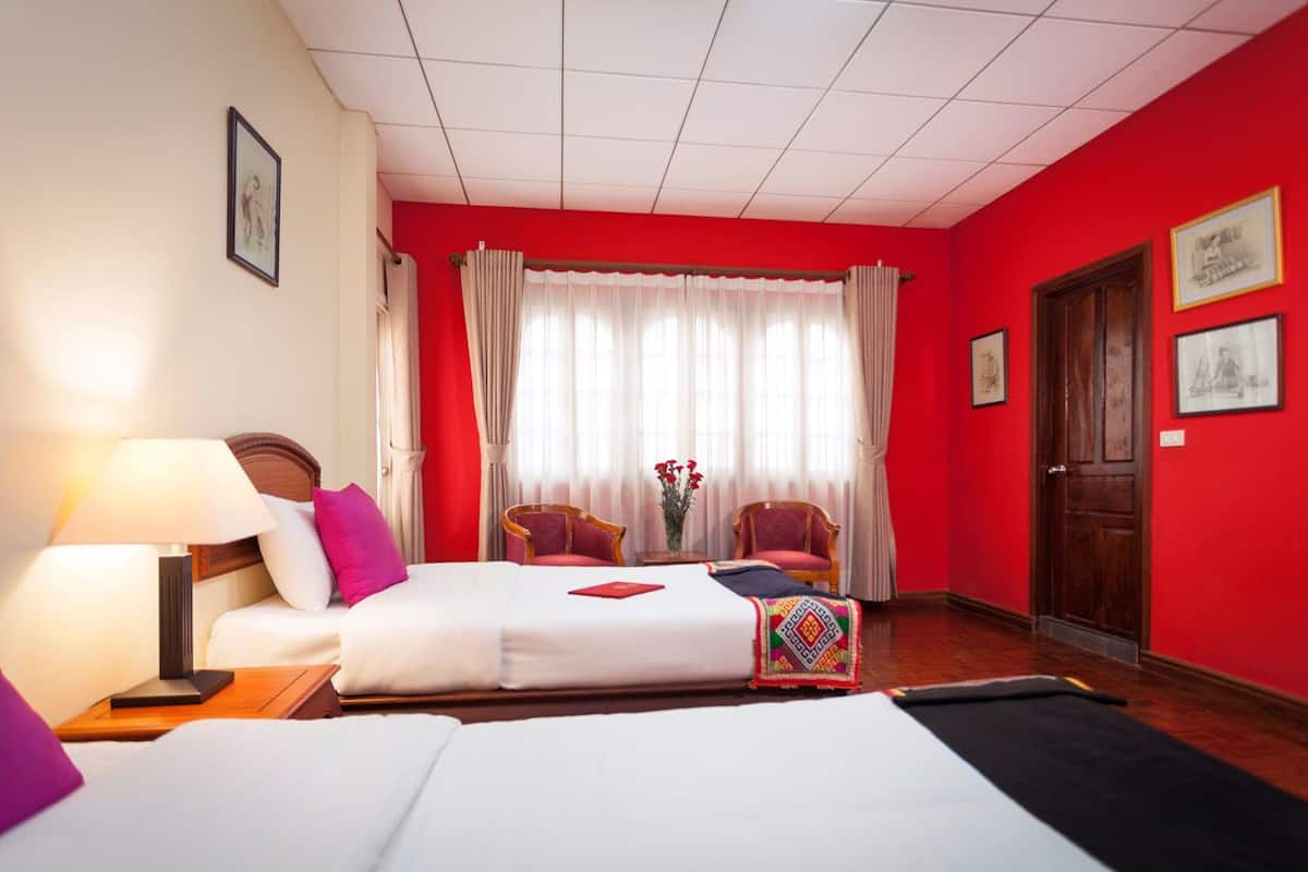 Habitación de hotel con dos camas, una pared roja, una ventana con cortinas blancas y una puerta.