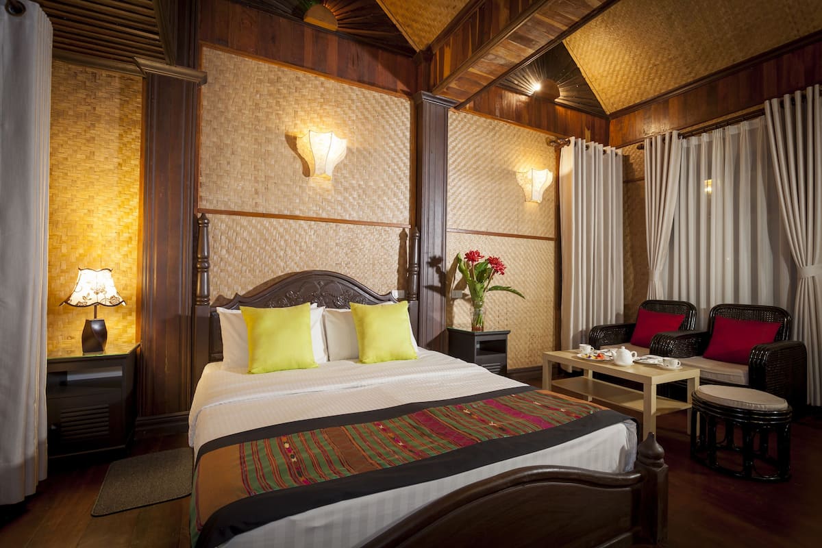 Habitación de hotel con una cama grande, una mesita de noche con lámpara, una pequeña mesa con tetera, una silla y un sofá con cojines rojos.