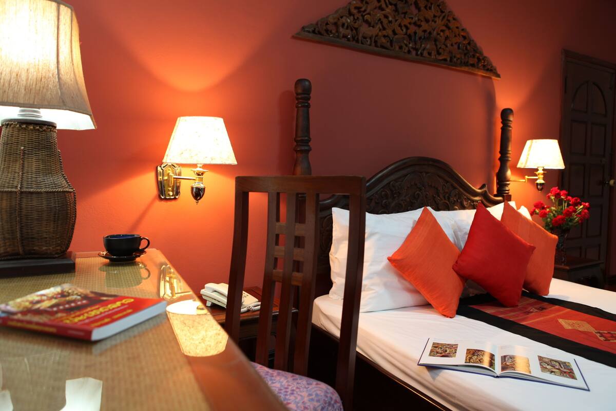 Habitación de hotel con cama, dos lámparas de noche, una silla de madera, una mesa con lámpara, un libro y un florero con flores.