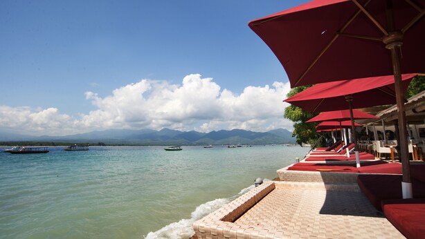 A beachside area with red umbrellas, a tiled walkway, and boats on the water. - Bel Air Resort & Spa (Gili Air)