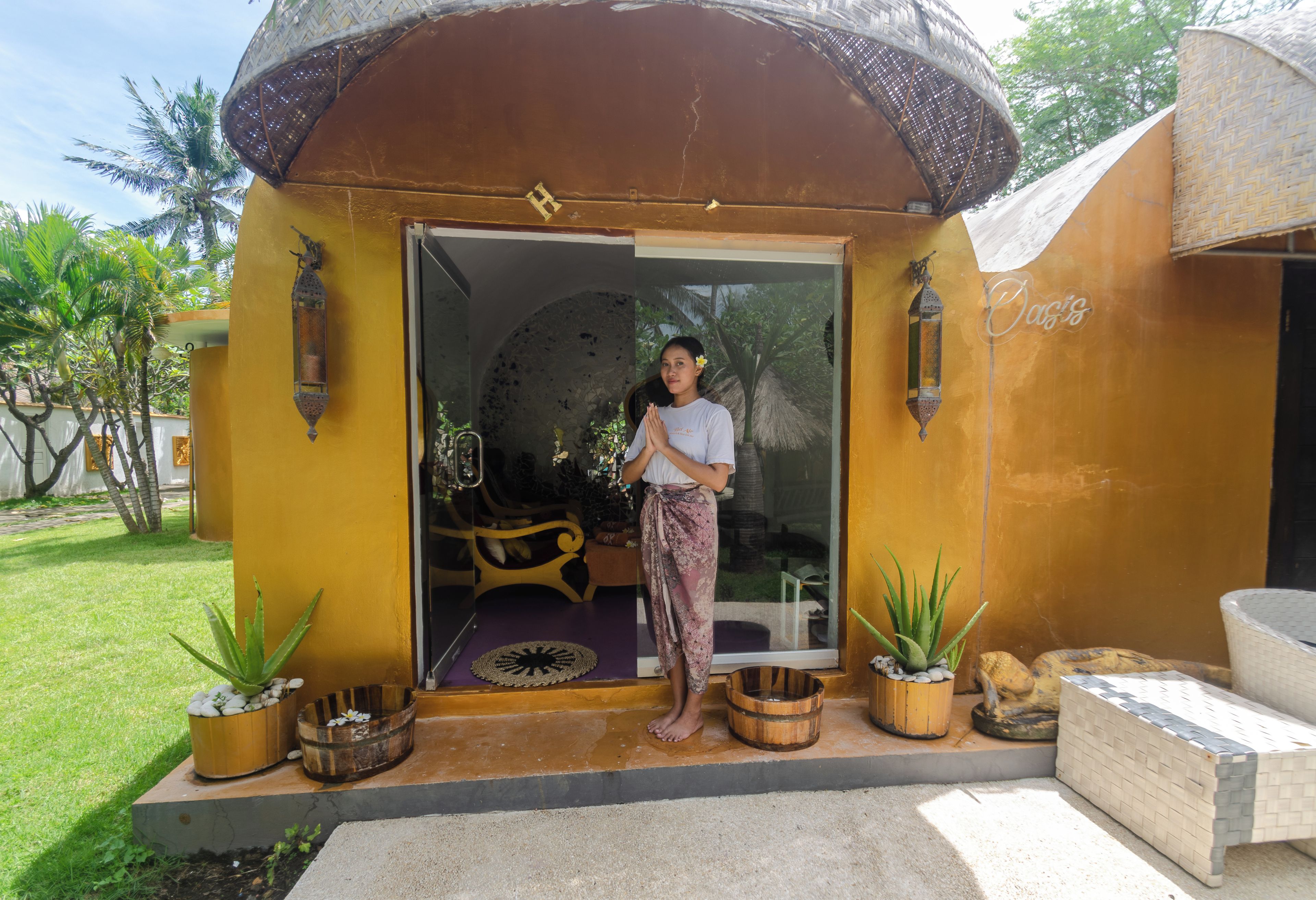 A person standing at the entrance of a yellow building with a thatched roof, a glass door, and a small garden area with potted plants.