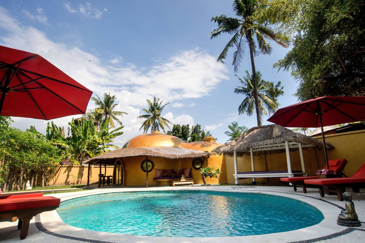 A swimming pool with red umbrellas, lounge chairs, and thatched-roof structures.