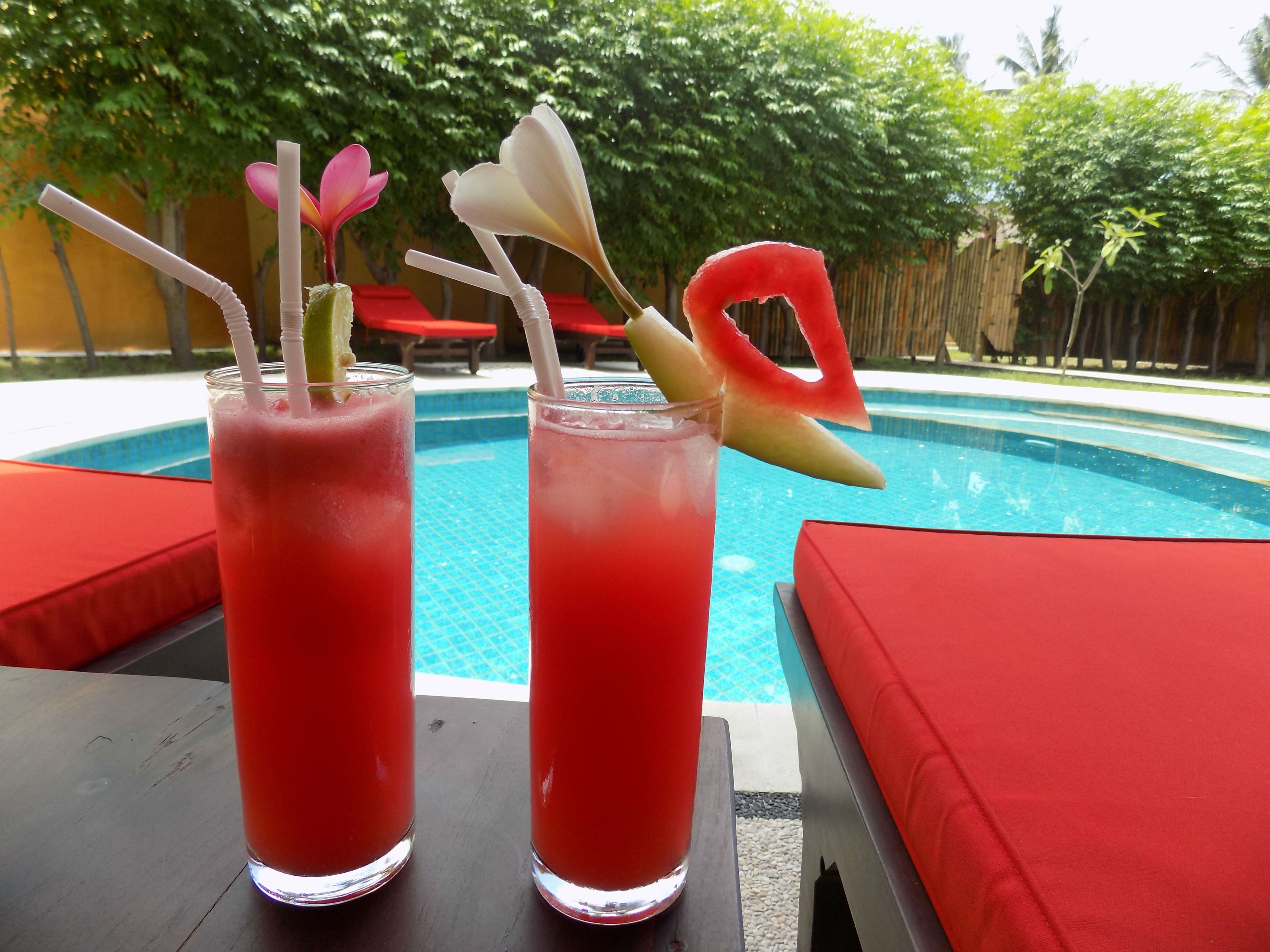 Dos vasos altos de bebida roja con pajitas y guarnición de frutas sobre una mesa junto a una piscina con sillas de descanso rojas.