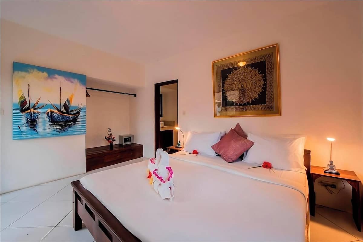 A hotel room with a bed, a painting of boats, a decorative mirror, and a framed artwork on the wall.