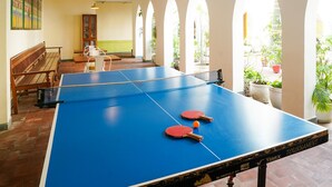 Ein Tischtennis-Tisch mit zwei Schlägern und einem Ball, eine hölzerne Bank und ein Bücherregal in einem Raum mit Rundbogenfenstern und hängenden Pendelleuchten.