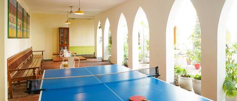 A ping pong table with two paddles and a ball, a wooden bench, and a bookshelf in a room with arched windows and hanging pendant lights.