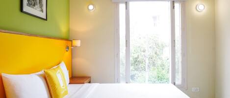 A hotel room with a bed, a nightstand, a picture on the wall, and an air conditioning unit above the bed.