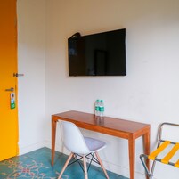 A small room with a wooden desk, a white chair, a television mounted on the wall, and a pair of water bottles on the desk.