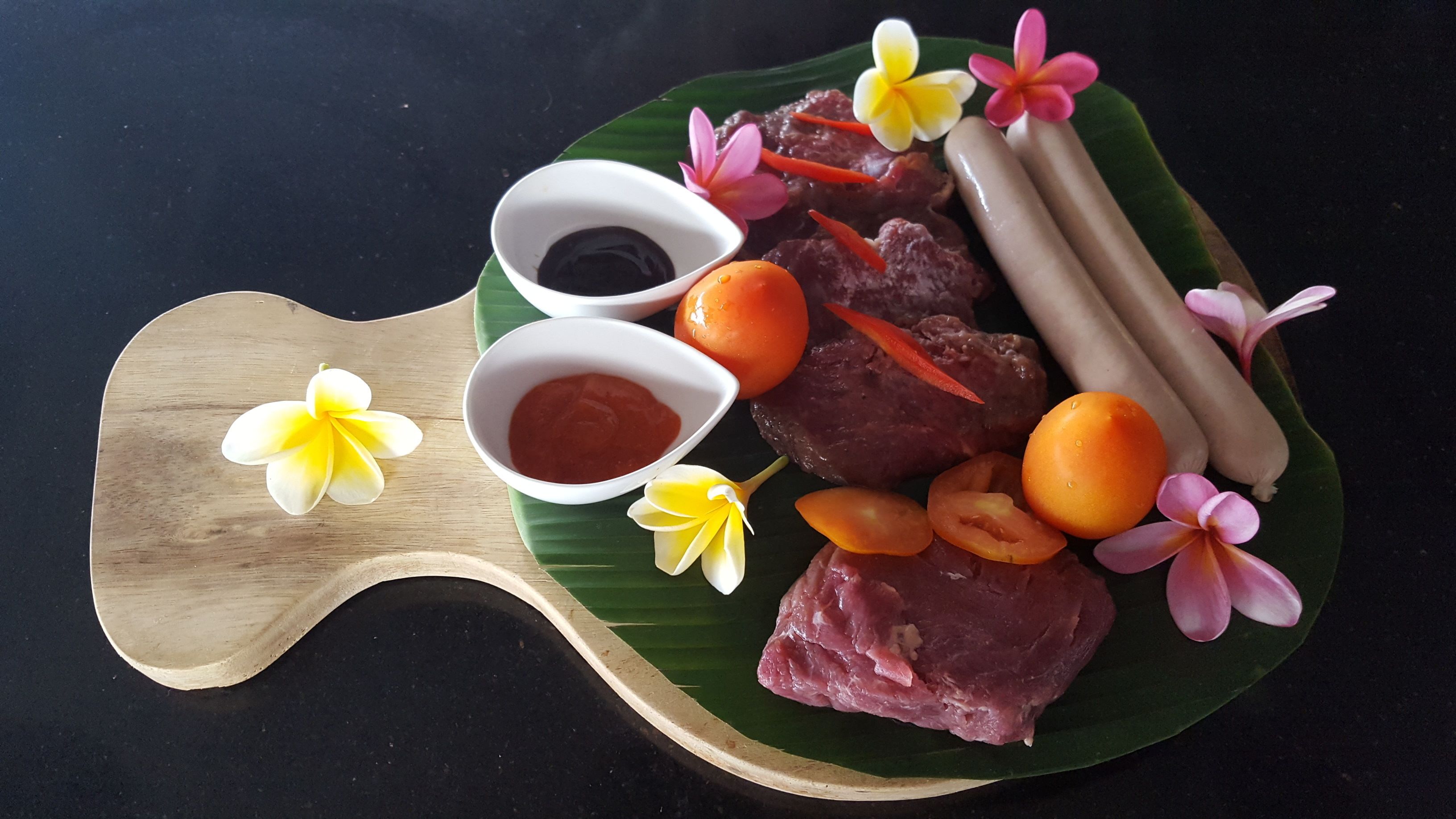 A wooden serving board with a green leaf, containing slices of meat, two whole oranges, and two small bowls of sauce.